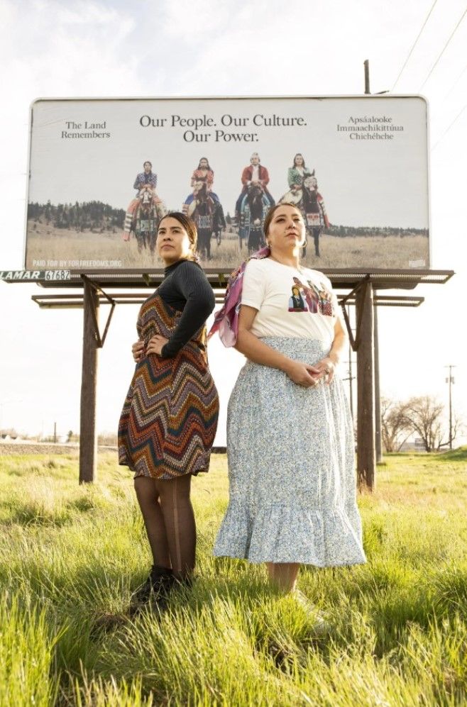 Photo by Robyn Iron. JoRee LaFrance (left) and Bethany Yellowtail pose in front of a billboard erected in west Billings as part of a national initiative by For Freedoms titled “Another Justice: By Any Medium Necessary.”