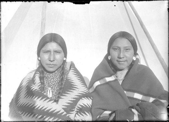 Clara White Hip / Iispaache (left) and Lottie Shell Bird stand in front of Crow teepees in 1886. / Historic photo from the Fr